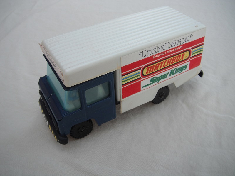 1. Dark blue cab/white plastic body/metal base/black plastic wheels/light blue windows/cream interior/dark blue steering wheel/labels w/various adverts of Matchbox ranges.